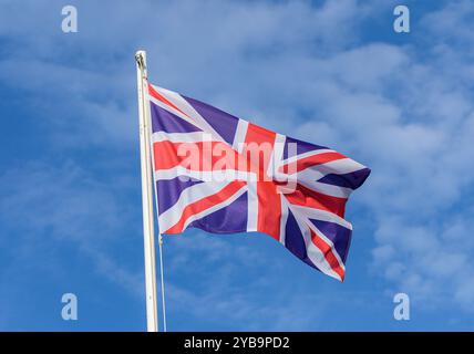 Der Union Jack flattert im sanften Wind und zeigt seine kräftigen Farben vor dem Hintergrund eines hellen, wolkengesprenkelten Himmels. Ein wahres Emblem von Britis Stockfoto