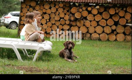 Kleine Jungen und Mädchen Kinder ruhen sich aus und spielen mit Hund im Hof des Landhauses. Freundschaftsbegriff, glückliche Kindheit. Deutsch Kurzhaarzeiger. Stockfoto