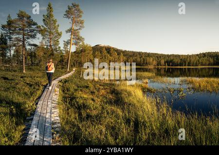 Frau spaziert entlang einer hölzernen Promenade am friedlichen See und genießt das warme Glühen der goldenen Sommerstunde mit den Skipisten von Idre im Hintergrund Stockfoto