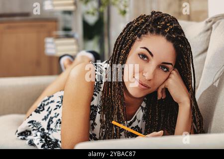 Eine junge Frau mit langen Zöpfen schreibt in einem Notizbuch auf ihrem Sofa und sieht entspannt und zufrieden in ihrem gemütlichen Wohnzimmer aus. Sie lächelt, verlor in Gedanken und cr Stockfoto