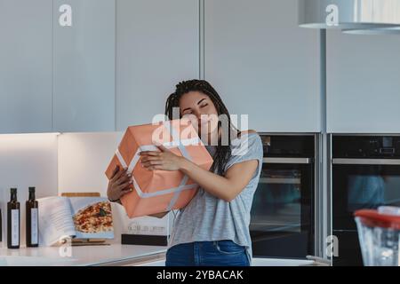 Die junge Frau umarmt eine Geschenkbox in ihrer gemütlichen Küche, die Augen geschlossen und strahlend lächelnd. Die Szene fängt Überraschungen und Feiern in einem modernen, leichten fi ein Stockfoto