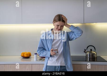 Depressive Frau trinkt Wasser, nachdem sie Pillen gegen Kopfschmerzen eingenommen hat, und versucht, mit Schmerzen in der Küche fertig zu werden Stockfoto