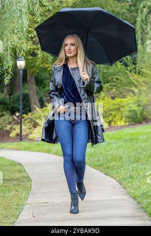 Eine atemberaubende Frau schlendert durch den Park, ihre Eleganz wird von einem schwarzen Regenschirm unterstrichen, inmitten der sich abzeichnenden Dunkelheit, bevor Regen die Erde küsst Stockfoto