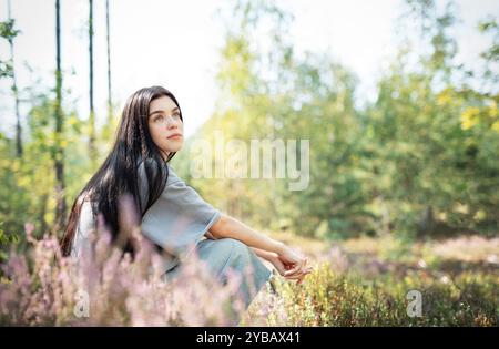 Eine junge Frau sitzt ruhig auf dem Waldboden, ihr langes Haar zieht sich über den Rücken. Sie erscheint tief in Gedanken, umgeben von blühenden Wildblumen Stockfoto