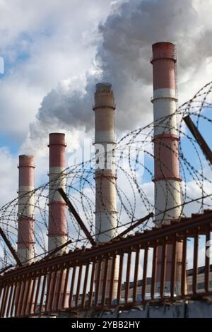 Dicker Rauch und Dampf weht aus den Fabrikschornsteinen hinter einem Stacheldrahtzaun Stockfoto