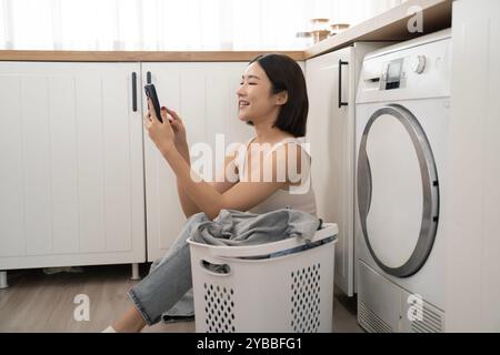 Eine junge Asiatin sitzt auf dem Boden neben einem Wäschekorb mit ihrem Telefon Stockfoto