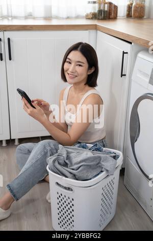 Eine junge asiatische Frau sitzt auf dem Boden neben einem Wäschekorb und benutzt ihr Telefon während ihrer Wäschearbeit Stockfoto