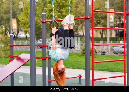 Frau, die kopfüber von den Fitnessringen auf dem Spielplatz hängt und Sport genießt. Stockfoto