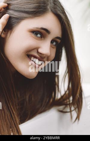 Eine junge Frau mit fließenden braunen Haaren lächelt warm und berührt ihr Haar sanft. Sie strahlt Glück und natürliche Schönheit aus, strahlt Jugendlichkeit und Wohlbefinden aus Stockfoto