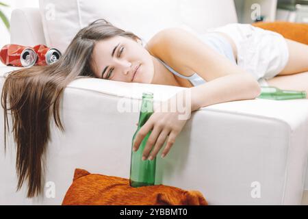 Müde Frau schläft nach einer Party auf einem weißen Sofa, umgeben von leeren Bierdosen. Ihr langes Haar fließt in ihr Kleid und zeigt die Gefahren von Alkohos Stockfoto