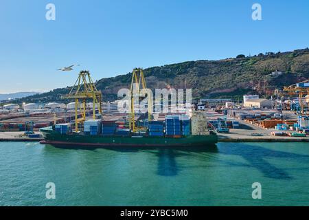 Barcelona. Espain - 28. September 2024: Ein mit Containern beladenes Frachtschiff, das am belebten Hafen von Barcelona angedockt wird, eingerahmt von großen Kränen und Clear blu Stockfoto