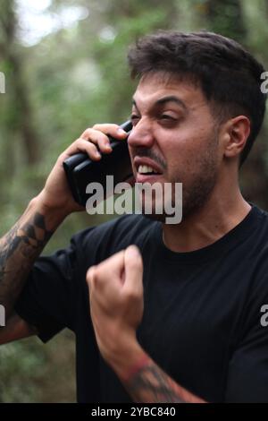 Ein frustrierter junger Mann schreit während eines intensiven Telefonanrufs in sein Handy und steht inmitten eines friedlichen Waldes Stockfoto