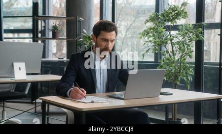 Fokussierter erwachsener Geschäftsmann arbeitet an einem Projekt im Büro mit Laptop, um Notizen zu schreiben, weißer Mann Geschäftsführer professioneller Mitarbeiter analysiert Informationen Stockfoto