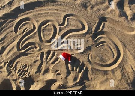 2025 das Jahr ist eine Zifferschrift auf dem Sand mit einem Manta claus Hut in Rot und weiß. Weihnachten Neujahrsfeier auf dem Meer. Stockfoto