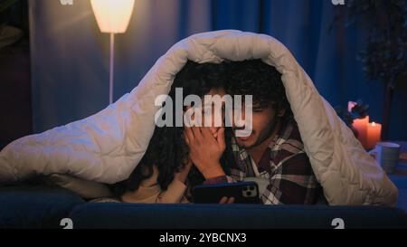 Gadget Internet-süchtig multirassisches Paar glückliche Familie Mann Frau Freund Freundin in der Nacht zu Hause unter der Decke, die ekelhafte schlechte Nachrichten Video Stockfoto