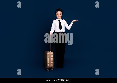 Foto in voller Größe von pensionierten weiblichen Flugzeugpiloten Koffer halten leeren Raum tragen Pilotenuniform isoliert auf dunkelblauem Hintergrund Stockfoto