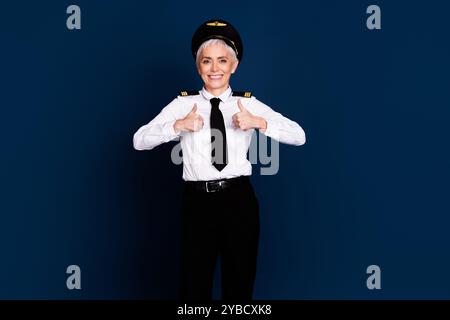 Foto-Porträt der hübschen pensionierten weiblichen professionellen Flugzeugpiloten Daumen oben tragen Pilotenuniform isoliert auf dunkelblauem Hintergrund Stockfoto