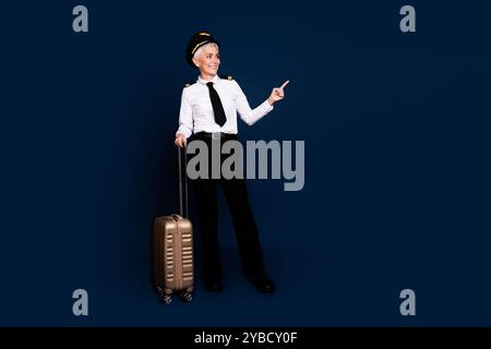 Foto in voller Länge von Rentnerin Dame kommerzieller Pilot Koffer Point Look Leere Space Tragen Pilotenuniform isoliert auf dunkelblauem Hintergrund Stockfoto