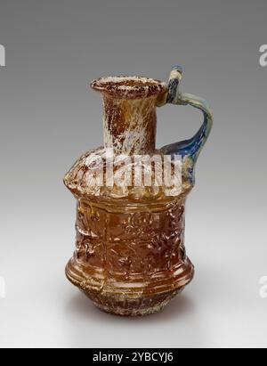 Oinochoe, 1. Jahrhundert n. Chr. zusätzliche Informationen: Diese kleine mundgeblasene Oinochoe besteht aus bernsteinfarbenem Glas und wurde in eine dreiteilige Form geblasen. Sein zylindrischer Körper ist mit einem Efefeufries zwischen Rippenmustern verziert. Die flache Basis ist mit konzentrischen Kreisen geprägt. Ein Griff mit einem Ellenbogen aus marmoriertem blauem Glas wurde auf die Schulter aufgebracht und bis zur Lippe gezogen. Diese Oinochoe ist in gutem Zustand. Die Oberfläche weist kleine Flecken mit schillernder und Lochfraß auf. Der obere Teil wurde mit großen Füllungen wieder an der Karosserie befestigt. Stockfoto