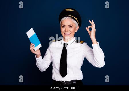 Foto von hübschen pensionierten weiblichen professionellen Flugzeugpiloten halten Tickets okey tragen Pilotenuniform isoliert auf dunkelblauem Hintergrund Stockfoto