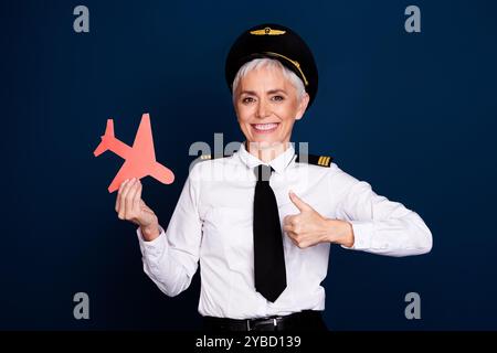 Foto von hübschen pensionierten weiblichen professionellen Flugzeugpiloten Papier Daumen oben tragen Fliegeruniform isoliert auf dunkelblauem Hintergrund Stockfoto