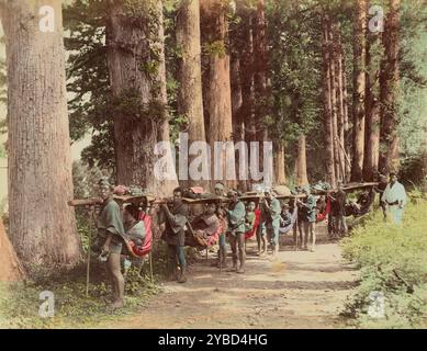 Kago Traveling Chair, 1870er-1890er Jahre Mehrere Männer tragen eine lange Stange, an der mehrere Stühle hängen. In jedem Stuhl sitzt eine Person. Die Männer stehen auf einem Feldweg in einem Wald. Stockfoto