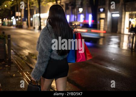 Lateinische Frau in modischer Kleidung wird von hinten gesehen, als sie nachts mit bunten Papiertaschen nach Hause geht. Die Stadt leuchtet Stockfoto