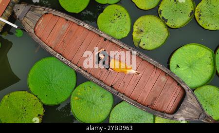 Asiatische Frau, die sich auf einem Langboot, umgeben von riesigen Victoria-Wasserlilien, im tropischen Regenwald von Phuket Thailand, entspannt Stockfoto