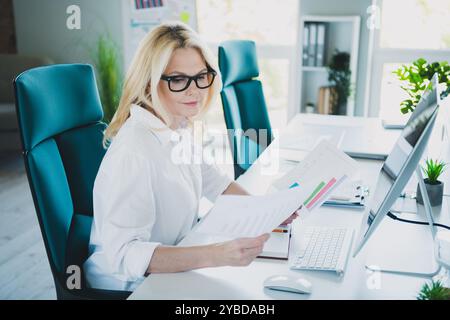 Foto einer blonden Arbeitgeberin mit Diagrammen, die ein formelles weißes Hemd tragen und Statistiken im Büro analysieren Stockfoto