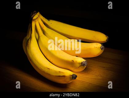 Ein Haufen von vier gelben Bananen, isoliert auf einem Tisch im Dunkeln Stockfoto