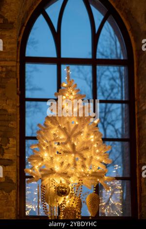 Ein weißer Weihnachtsbaum, der mit warmen Lichtern und Ornamenten geschmückt ist, ist vor einem großen Fenster sichtbar, wo Stockfoto