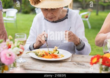 Ältere Frau mit Sonnenhut und weißem Pullover, die bunte, nahrhafte Mahlzeit am Tisch im Freien essen. Senior Lifestyle Bild zeigt gesunde Ernährung, soziale Stockfoto