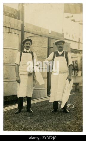 Anfang der 1900er Jahre Postkarte von zwei Arbeitern in Schürzen, möglicherweise Mühlen, neben großen Metallfässern, die wahrscheinlich Behälter für Mais und Futtermittel waren. Ich dachte, es wäre Talgarth Mill. Die Männer stehen stolz darauf, dass sie fotografiert werden, vielleicht die neuen Mieter der Mühle. Die Fässer warten auf die Installation. Ein neues Unternehmen. Die patriotische Union Jack könnte darauf hinweisen, dass dies während des 1. Weltkriegs um 1914 erfolgte. Von O. Jackson, Wellington Studio, Talgarth, Powys, Wales. Um 1910 Stockfoto