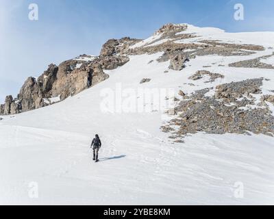 Ein einsamer Wanderer klettert einen schneebedeckten Hang hinauf zu einem felsigen Gipfel in Briancoon, Frankreich. Die weite, zerklüftete Landschaft wird vom klaren blauen Himmel beleuchtet Stockfoto