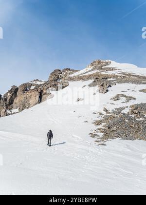 Ein einsamer Wanderer klettert einen schneebedeckten Hang hinauf zu einem felsigen Gipfel in Briancoon, Frankreich. Die weite, zerklüftete Landschaft wird vom klaren blauen Himmel beleuchtet Stockfoto