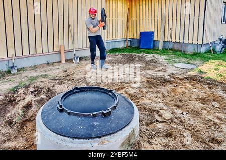 Ein neuer Klärgruben aus Betonringen wird auf dem Hausgelände im ländlichen Raum installiert, der Arbeiter trägt eine Mannlochabdeckung, um sie gut abzudecken. Stockfoto