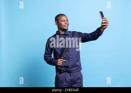 Seriöser Sicherheitsbeamter macht gerne Fotos mit seinem Schlagstock vor blauem Hintergrund und posiert mit einer Waffe auf der Handykamera. Ein zuversichtlicher Agent macht Fotos im diensthabenden Studio. Stockfoto