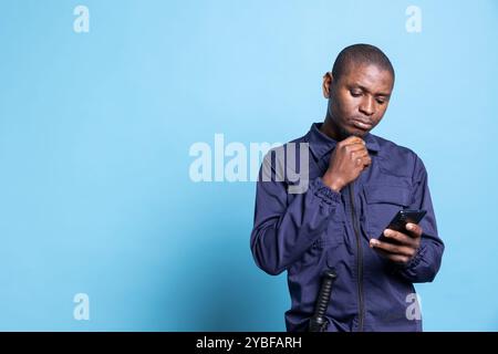 afroamerikanischer Sicherheitsbeamter navigiert im Online-Browser am Telefon und sendet Nachrichten in seiner Arbeitsunterbrechung. Agent in Uniform prüft Social-Media-Konten und Apps vor blauem Hintergrund. Stockfoto
