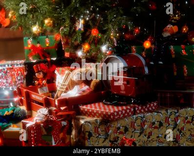 Unter einem Weihnachtsbaum, der mit Ornamenten und Lichtern verziert ist, befindet sich eine Sammlung eingewickelter Geschenke. Ein roter Spielzeugzug, ein Teddybär mit Trompete und ein Stockfoto