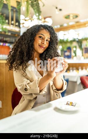 Vertikales Porträt einer lateinischen Schönheitsfrau, die warmen Kaffee genießt, in einer modernen Cafeteria sitzt und in die Kamera lächelt Stockfoto