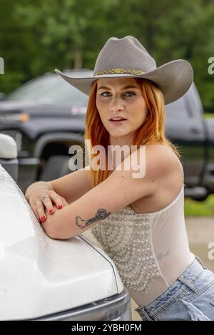 Ein wunderschönes rothaariges Model posiert in einer ländlichen Umgebung und trägt Cowgirl-Kleidung an ihrem Truck Stockfoto