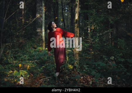 Eine Frau in einem roten Pullover steht anmutig in einem lebendigen Wald und genießt das Sonnenlicht, das durch die Bäume filtert, während sie die umliegenden Foli beobachtet Stockfoto