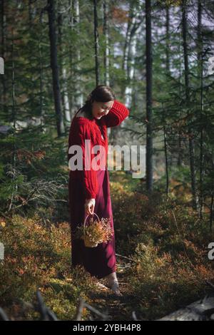 Eine Frau in einem gemütlichen roten Pullover schlendert im Herbst durch einen ruhigen Waldweg und hält einen Korb voller leuchtender Blumen in der Farbe Stockfoto