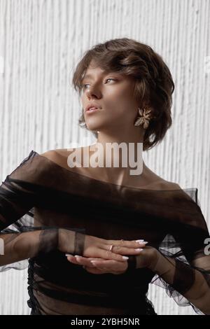 Eine Frau strahlt Eleganz aus in einem transparenten schwarzen Kleid, die eine nachdenkliche Pose zeigt. Der strukturierte weiße Hintergrund unterstreicht die moderne Mode-Ästhetik, Kreation Stockfoto