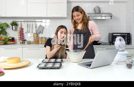 Zwei junge Frauen auf einer Schürze bereiten in der Küche Nachtisch zu, während sie auf dem Laptop im Online-Rezeptbuch nachschauen. Stockfoto