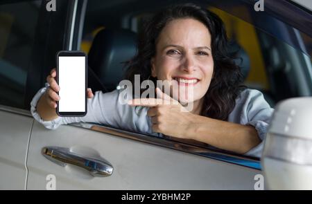 Die weiße Frau sitzt mit dem Fenster nach unten auf dem Fahrersitz eines Autos. Sie lächelt und zeigt auf das Smartphone mit einem leeren Bildschirm in Richtung Stockfoto
