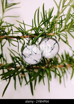 Handgefertigte Harz runde Marmor Textur Stein Ohrringe transparente Oberfläche am grünen Rosmarinzweig. Naturinspirierter Schmuck, handgefertigter Silberschmuck Stockfoto