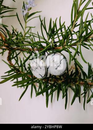 Handgefertigte Harz runde Marmor Textur Stein Ohrringe transparente Oberfläche am grünen Rosmarinzweig. Naturinspirierter Schmuck, handgefertigter Silberschmuck Stockfoto