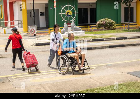 Eine Frau, die einem älteren Mann im Rollstuhl hilft, die Straße in der Marine Parade Promenade Singapur zu überqueren Stockfoto