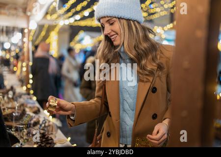 Junge Frau in Winterkleidung, die den Weihnachtsmarkt besucht. Lächelnde Kundin mit Geschenkbox, umgeben von festlichen Lichtern und Dekorationen Stockfoto